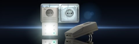 A lisa device plugged into an outlet and a rectangular device lying in front of the outlet with two inputs and a plug on the back. 