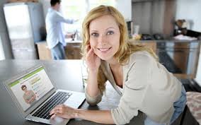 Photograph of a woman leaning over a table with her left hand on the keyboard of a laptop and her right hand on the side of her face, which is looking straight ahead.