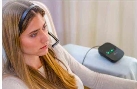 A young lady is sitting in an armchair wearing a headband-type device with an arm extending out from the ear toward the mouth. A small rectangular corded device with 2 green lights is next to her on the chair's arm.