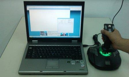 A silver laptop open to a blue desktop with several open windows. To the right, a user operates a hand joystick that is placed on the desk.