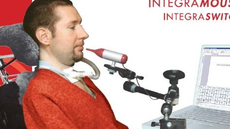 The wine bottle-shaped mouthpiece is attached to a black mounting arm positioned in front of a user sitting in a wheelchair in front of a computer.