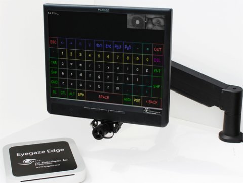 A small, square device next to a desktop screen with a camera at its base on an adjustable, vertical mount. 