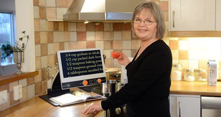 The FHD 13 on a countertop. A woman magnifies the recipe below the monitor, projecting it in white text with a black backround.