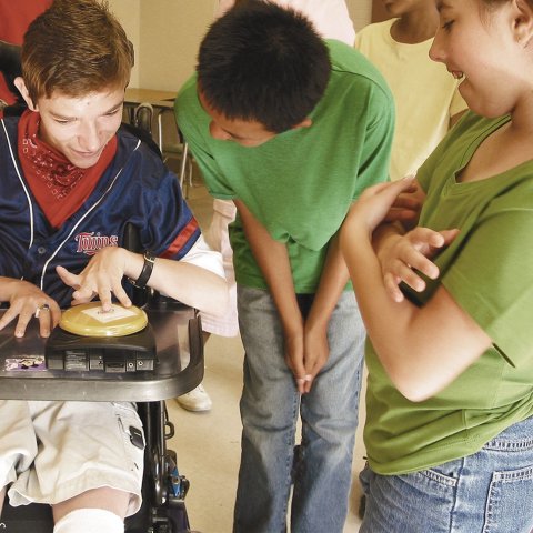 User in wheelchair with left hand on yellow switch and bystanders watching.