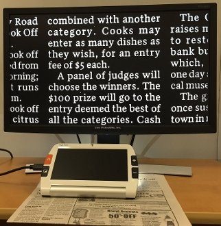 Magnifier with HDMI output plugged into left side. Magnifier is placed over a newspaper article with light shining to highlight the area being viewed. Magnified section of article displayed on LCD screen with white letters and black background.
