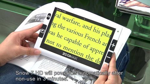 Camera/magnifier screen the size of a small book is held in two hands. The device screen is displaying printed text using a high-contrast filter with a yellow background.