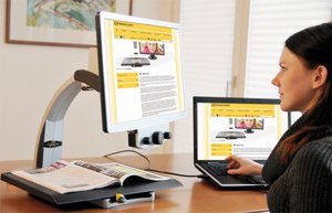Medium-sized monitor with a connected platform underneath at its base. The platform allows materials to be placed and magnified. On the bottom front edge of the monitor, there are three round knobs. A laptop is connected and both device screens are displaying the same material.