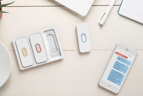 An iPhone with an audio transcription displayed. Next to the phone, there are four wearable microphones, which are long, white, and oval/rectangular, and have colored ovals in the center.