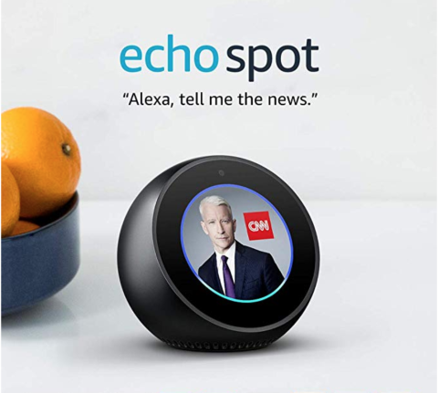 A small, black, and round device roughly the size and shape of a small alarm clock, featuring an LCD display screen. A quote reads, "Alexa, tell me the news."
