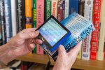 One hand of an individual holding the Maggie magnifier over a portion of a book which is held in the other hand of the individual, The Maggie has a white screen showing magnified wording from the book.