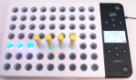 A 9x7 grid of peg holes in a whiteboard that has a cord attached on the upper right side. Seven of the holes have a neon blue light that is on, and 4 of those holes have long yellow pegs in them. The right side of the board has a narrow strip which is black and has two sets of buttons, each set for a person sitting on either side of the board. These buttons are labeled with directional arrows. There is also a speaker on both sides, and a button in each of the two corners labeled "start/stop".