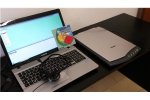 Laptop on a table with a Kit Ti Guido CD case leaning against the monitor; a headset rests on the keyboard and a flat, rectangular scanner sits to the right.