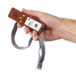 Hand holding a long, brown rectangular device with a speaker on one end and three yellow buttons underneath attached to a lanyard.