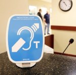 Blue and white device sitting on counter in office setting. Device features an image of an outline of an ear with an arrow diagonally through it and a small capital letter T on the lower right.