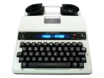A TTY device resembling an electric typewriter. The keys and display panel are black, while the device body is off-white.