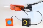 A rectangular orange plastic device with a clear plastic point connected to a flexible arm. Two small square devices are attached with round buttons.